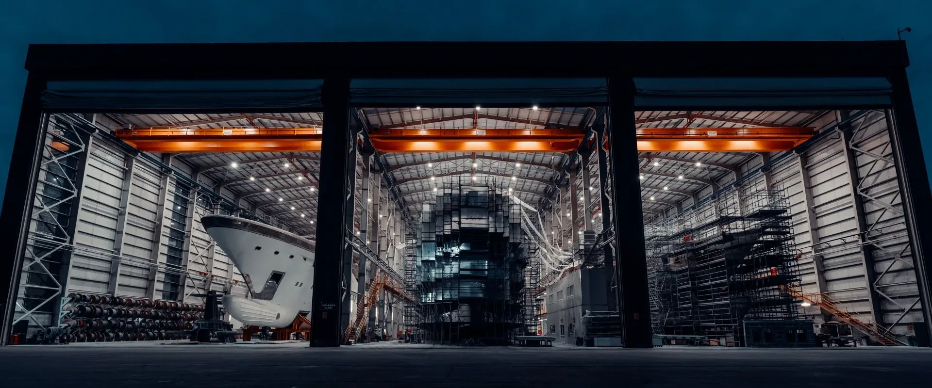 Bilgin Yachts build halls at dusk, doors open with a hull and scaffolding inside under lit gantry cranes