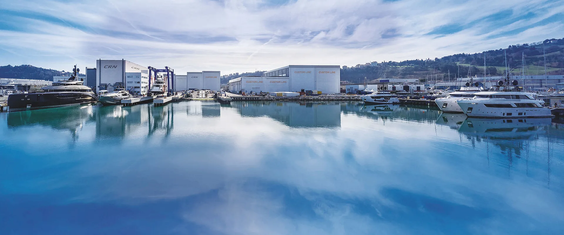 The Ancona superyacht yard — CRN and Custom Line build halls mirrored in still harbour water, with yachts along the quays